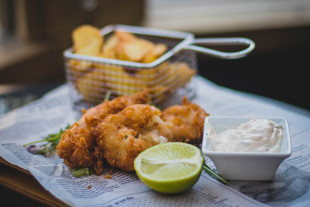 Plato de pescado frito crujiente servido con una rodaja de limón y una salsa blanca en un pequeño recipiente, acompañado de papas fritas en una cesta de metal