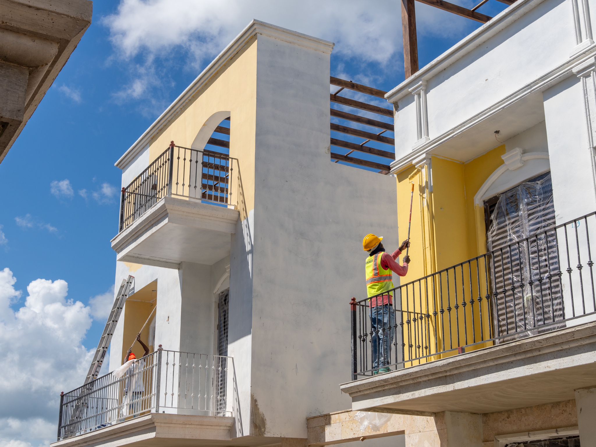 Trabajador pintando balcones en la fachada de un edificio en construcción en Pueblito Caribeño, con arquitectura de estilo colonial y cielo despejado de fondo